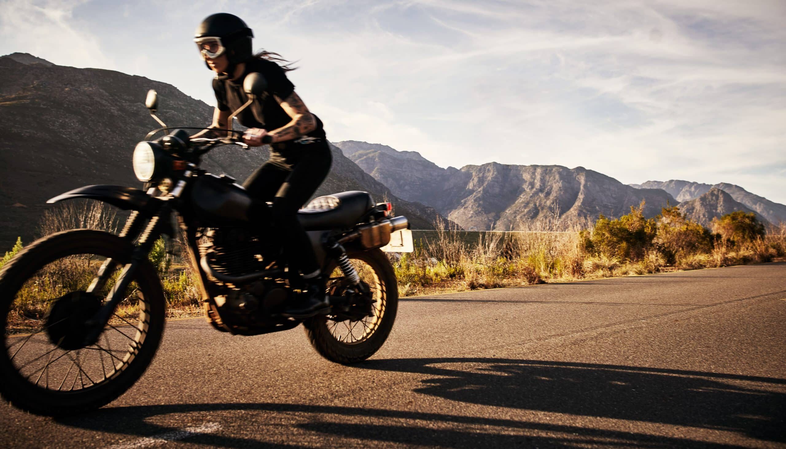 A motorcyclist wearing protective gear rides a black motorcycle on an open road with mountains in the background.