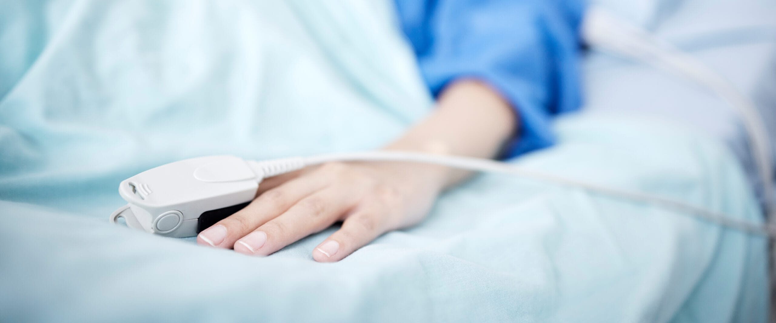 Close-up of a person's hand resting on a hospital bed, wearing a heart rate monitor, with the person lying in the background covered by a blue blanket.
