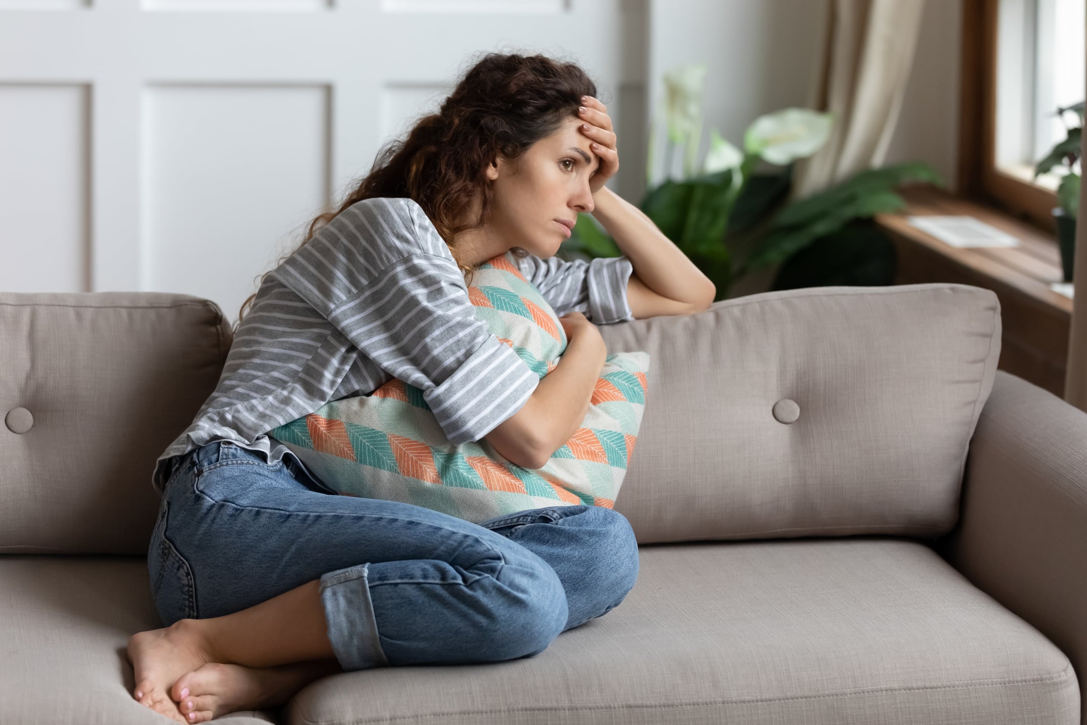 A woman sits on a couch, hugging a pillow, looking distressed, with her hand resting on her forehead.