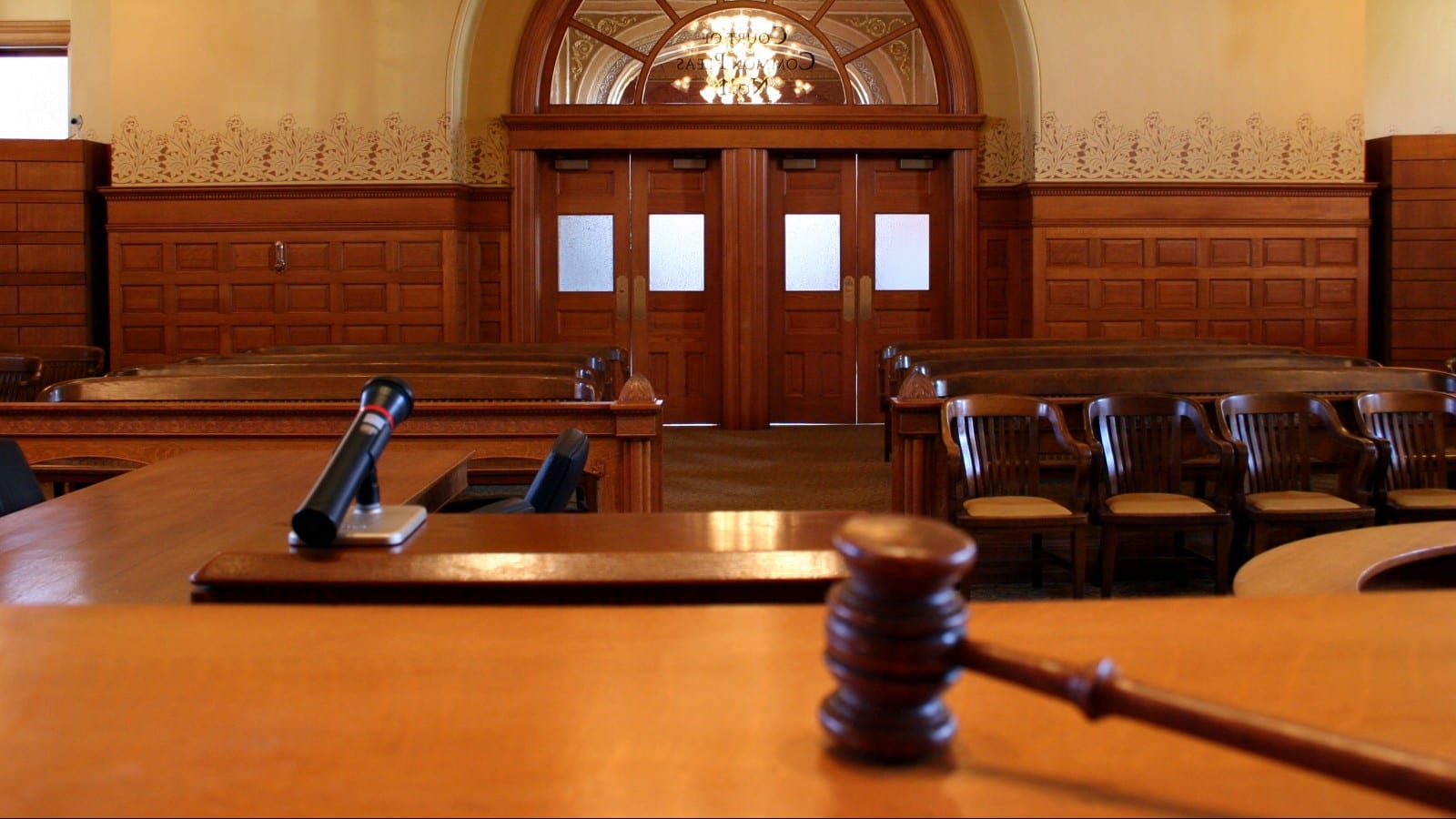 A wooden gavel rests on a judge’s bench in an empty courtroom, with a microphone and rows of chairs visible.