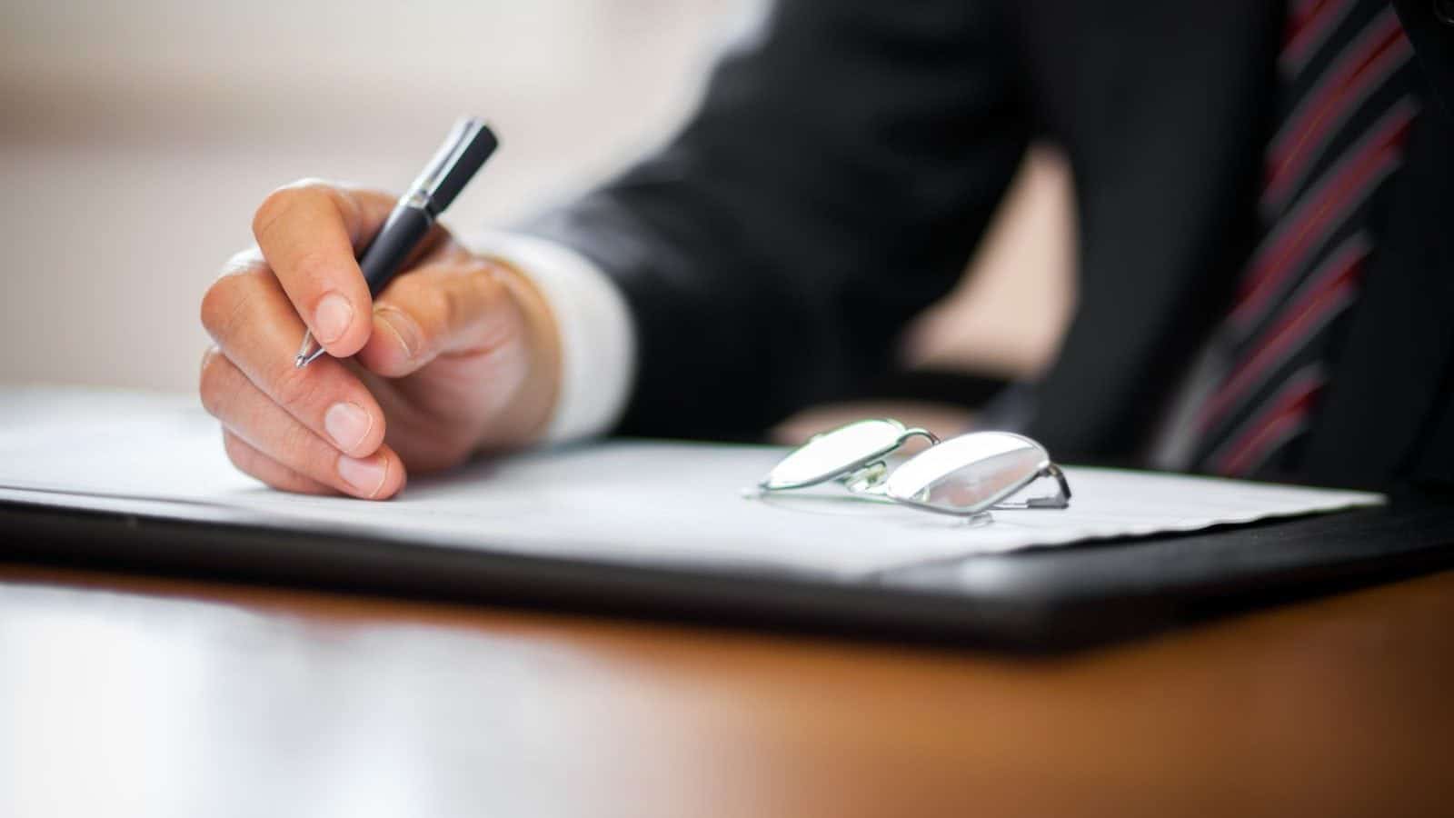 Close-up of a business professional in a suit writing on a document with a pen, with eyeglasses resting on a clipboard on a wooden desk.