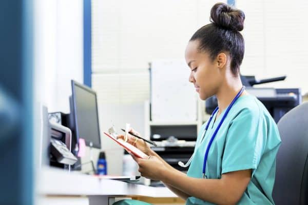 A nurse in green scrubs sits at a desk in a medical office, reviewing a clipboard.