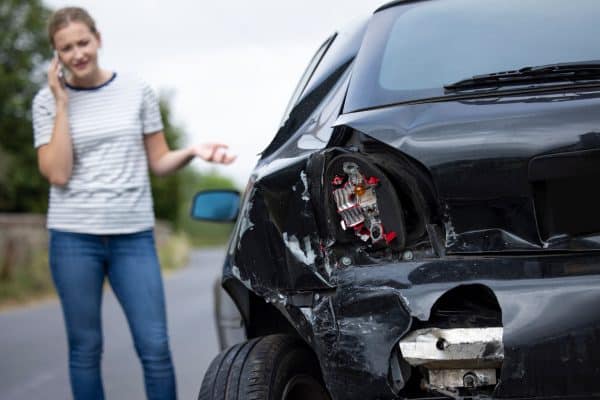 A woman stands beside her car, which has significant rear-end damage, while speaking on the phone and looking concerned.