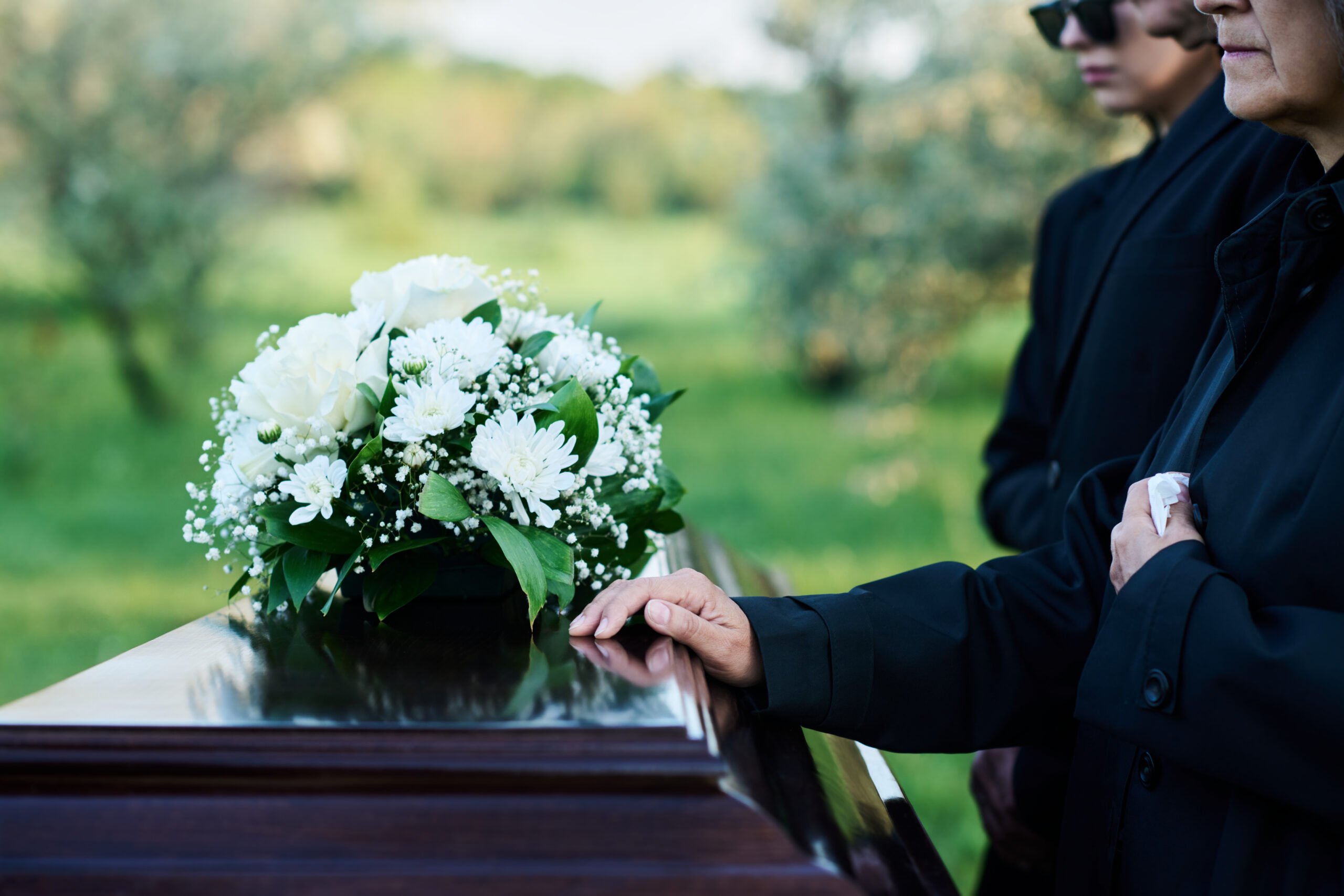 A close-up of mourners standing by a casket adorned with white flowers during a funeral.