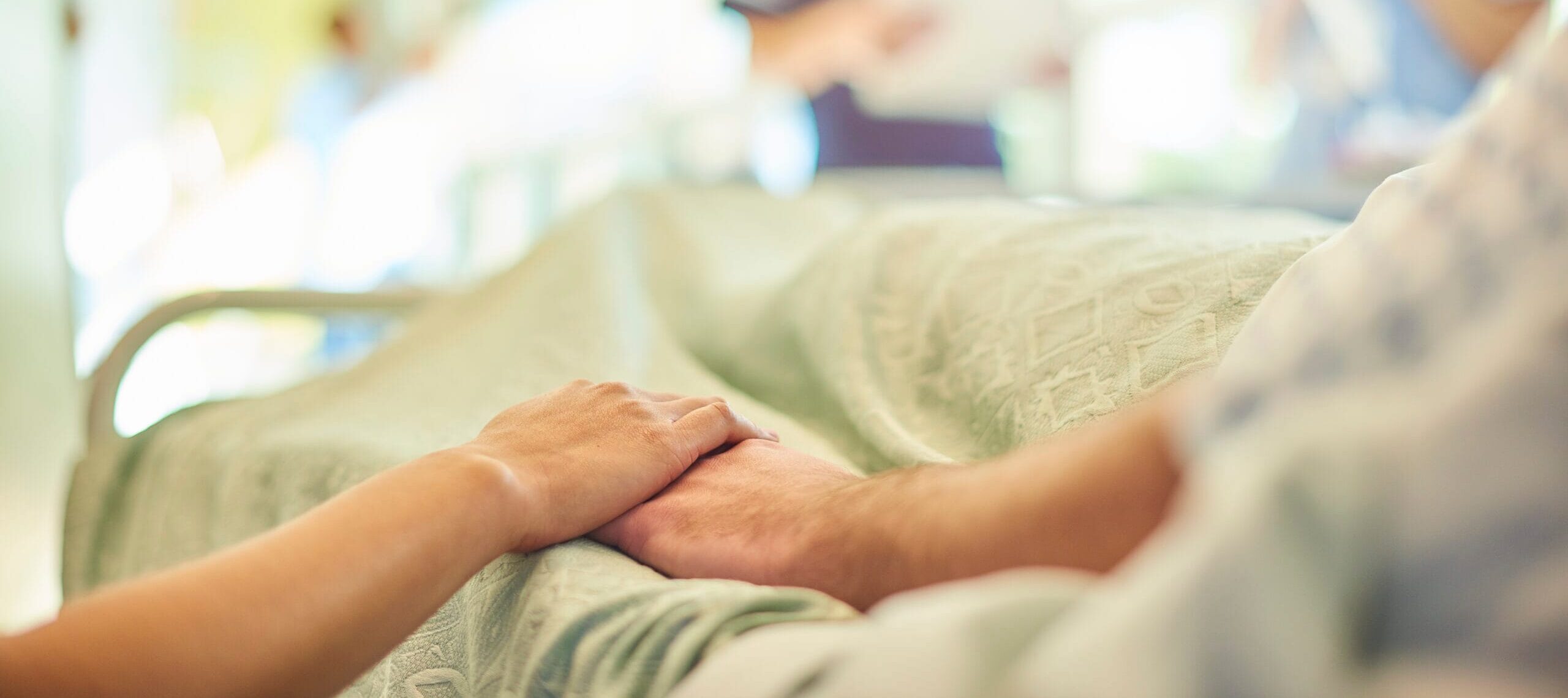Close-up of a patient lying in a hospital bed holding hands with a loved one, with two medical professionals in the background.